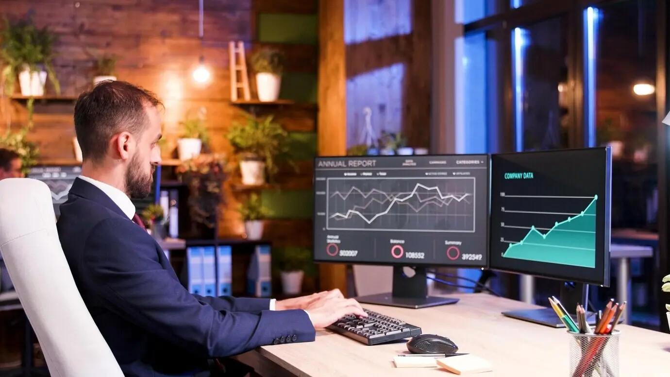 Менеджер поздно ночью работает с финансовыми данными на двух мониторах.